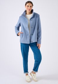Chaqueta de mezcla de lana en azul claro con un cuello alto, cierre de botones y bolsillos laterales, combinada con pantalones de mezclilla azules y zapatillas blancas.