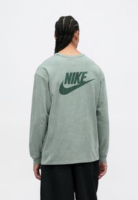 Tricou cu mânecă lungă, verde, de la Nike, dintr-un amestec de bumbac, cu o croială lejeră, având un logo mare de culoare verde închis pe spate, purtat cu pantaloni negri.