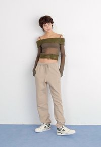 Olivgrünes, schulterfreies Top mit gerippter Textur, kombiniert mit beigen Sweatpants. Das Model trägt weiße Sneakers mit olivfarbenen Akzenten.