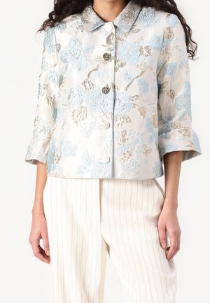 Femme portant une veste courte texturée avec des motifs floraux bleus et argentés et un pantalon blanc perle à fines rayures.