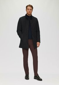 Schwarzer Mantel mit Knopfverschlüssen, hohem Kragen und seitlichen Taschen. Getragen über einem marineblauen Hemd und braunen Hosen, ergänzt durch schwarze Schuhe.