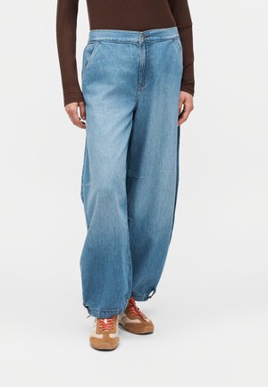 Osoba ubrana w jasnoniebieskie szerokie jeansy, brązowy prążkowany longsleeve oraz beżowe sneakersy z pomarańczowymi sznurówkami, stojąca na białym tle.