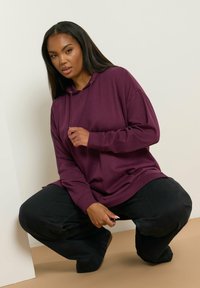Sudadera morada de gran tamaño con un bolsillo frontal y capucha con cordón, combinada con pantalones negros holgados, creando una silueta relajada.