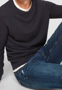 Granatowy sweter o fakturze z wypukłymi poziomymi liniami, z okrągłym dekoltem, noszony na białej koszuli. W zestawie z niebieskimi dżinsami z przetarciami.