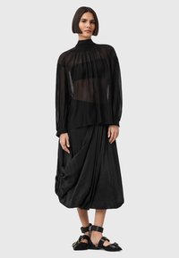 Blusa nera trasparente a maniche lunghe con scollo arricciato, abbinata a una gonna nera drappeggiata a vita alta e sandali neri bassi.
