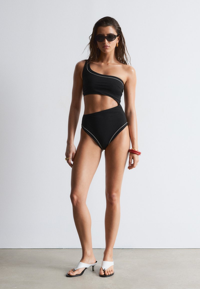Femme debout, portant un maillot de bain noir à une épaule avec découpes, des lunettes de soleil noires, des sandales blanches à talons et des bracelets rouges au poignet droit.