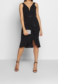 Svart klänning med djup V-ringning, åtsittande liv och volangsläpp. Har ett bälte i midjan och är parat med svarta klackar med remmar.