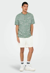 Camiseta verde de manga corta con un patrón floral, combinada con pantalones cortos blancos texturizados y zapatillas blancas con rayas negras en los calcetines.