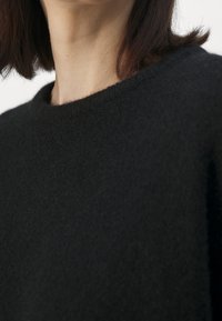 Pull noir avec un col rond, tissu doux et texturé, coupe légèrement oversize, mettant en valeur des détails de design minimalistes.