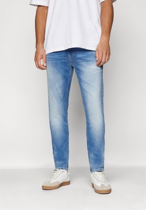 Mann trägt hellblaue ausgewaschene Jeans, weiße Sneakers und ein schlichtes weißes T-Shirt, steht vor einem einfarbigen Hintergrund.