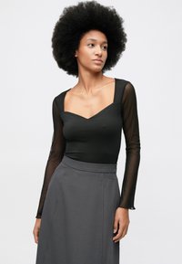 Moteris su natūraliu afro šukuosena, dėvinti juodą permatomą ilgomis rankovėmis viršutinę dalį ir pilkai aukštu juosmeniu sijoną, žiūri į šoną prieš baltą foną.