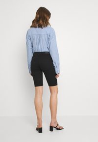 Černé džínové kraťasy s přiléhavým střihem, doplněné modrou pruhovanou košilí a černými sandály na podpatku. Modelka otočená zády k fotoaparátu.