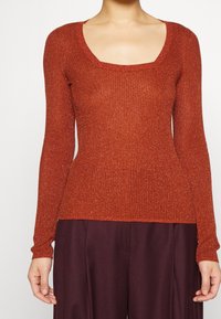 Femme portant un pull à manches longues côtelé de couleur rouille avec un décolleté carré, associé à un pantalon violet foncé.