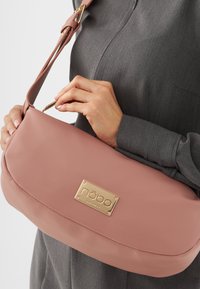Borsa a tracolla in pelle rosa con forma curva, caratterizzata da una placca logo in metallo dorato. La tracolla è regolabile e la texture è liscia.