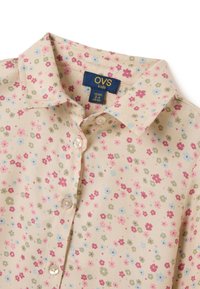 Camicetta beige chiaro con un motivo floreale in rosa, blu e verde. Presenta un colletto e cinque bottoni. Tessuto morbido al tatto.