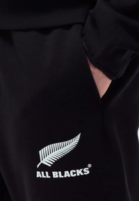 Crne trenirke sa bočnim džepom, s bijelim logotipom paprati i natpisom "ALL BLACKS" otisnutim blizu rubova. Glatka tekstura tkanine.