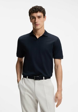 Junger Mann mit lockigem Haar, der ein schwarzes kurzärmeliges Poloshirt trägt, das in eine hellbeige Hose mit schwarzem Gürtel gesteckt ist, steht mit einer Hand in der Tasche.