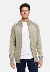 Beżowy kardigan na zamek błyskawiczny z prążkowanym kołnierzem i mankietami, dzianinowa tkanina o fakturze oraz kontrastujący zamek. Noszony na białej koszulce i granatowych dżinsach.