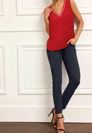 Červený top bez rukávů s krajkovými detaily na výstřihu, kombinovaný s tmavě modrými skinny džíny a černými sandály na vysokém podpatku s řemínky.