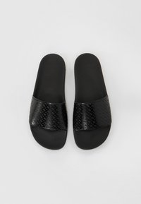 Sorte slip-on sandaler med en tekstureret sort rem, der har et geometrisk mønster. Sålen er flad og lavet af sort gummimateriale.