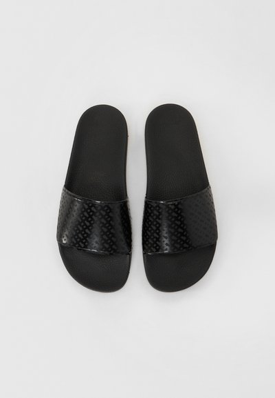 Sandales noires à enfiler avec une sangle noire texturée arborant un motif géométrique. La semelle est plate et fabriquée en caoutchouc noir.
