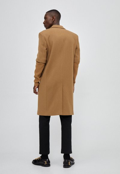 Long beige coat with a structured design, featuring ribbed sweater-like sleeves, paired with black trousers and black buckled shoes.