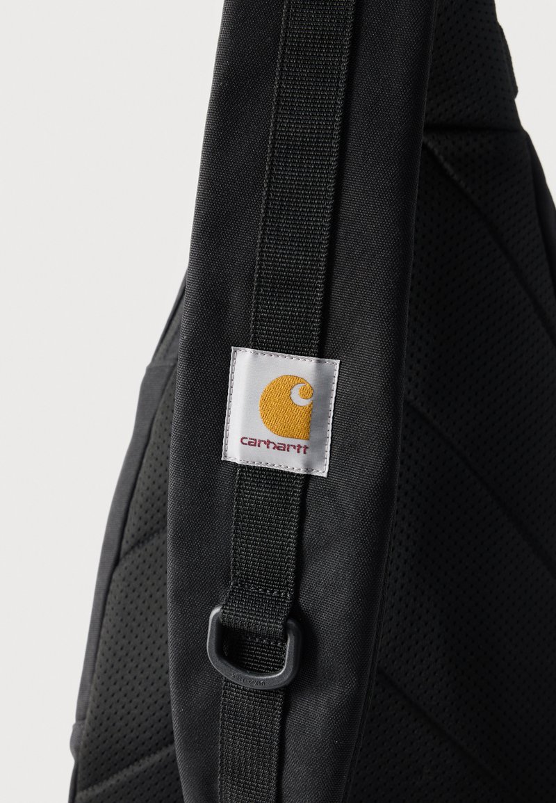 Correa negra de mochila con respaldo de malla acolchada y parche con el logo de Carhartt en costuras amarillas y rojas.