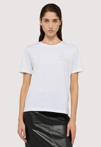 Camiseta de algodón blanca con mangas cortas, escote redondo y un logo impreso sutil en la parte superior izquierda. Textura suave, corte casual.