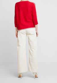 Pull en maille rouge à manches trois-quarts, associé à un pantalon large en velours côtelé crème. Chaussures à talon léger.