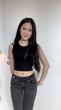 Junge Frau mit langen, dunklen Haaren, die ein schwarzes, ärmelloses Crop-Top und hochtaillierte dunkle Jeans trägt, steht vor einer weißen Tür.