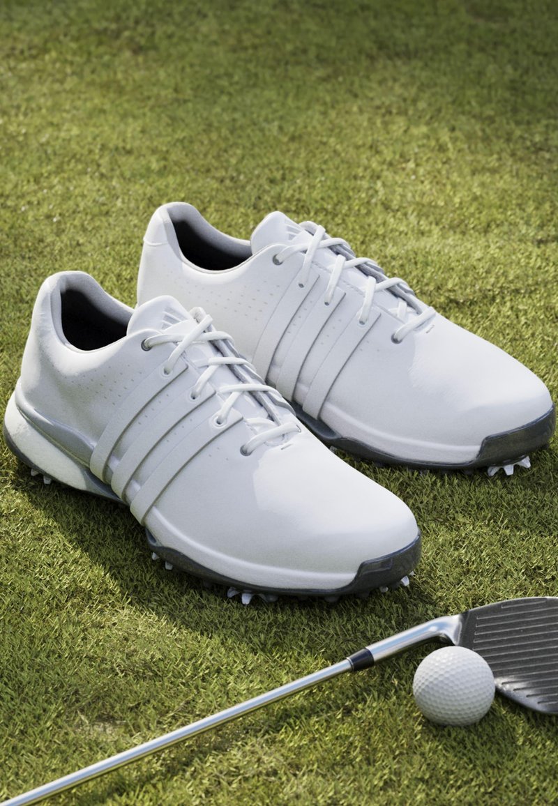 Bílé golfové boty s texturovaným syntetickým svrškem, šněrováním, třemi bočními pruhy a špičatou podrážkou na zelené trávě s golfovým klubem a míčem.