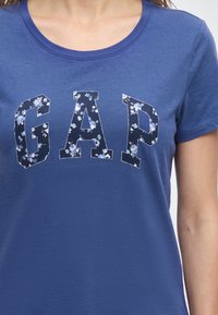 Femme portant un t-shirt bleu GAP avec un motif floral à l'intérieur de grandes lettres majuscules sur la poitrine.