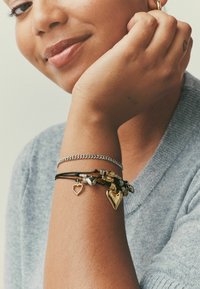 Sorte læder- og sølvkædearmbånd med gyldne hjertecharms; hånd hviler mod hagen, iført en grå sweater. Detaljerede teksturer synlige.