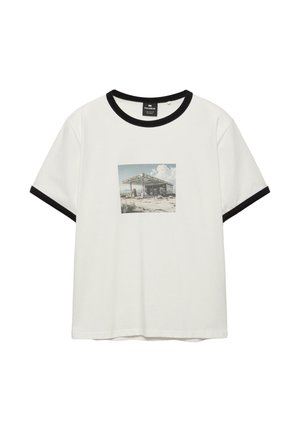 Wit T-shirt met korte mouwen en zwarte bies aan de kraag en mouwen, met een vervaagde afbeelding van een verlaten tankstation.