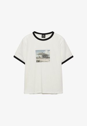 Wit T-shirt met korte mouwen en zwarte bies aan de kraag en mouwen, met een vervaagde afbeelding van een verlaten tankstation.