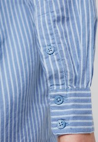 Camicia azzurra con strisce bianche verticali. I polsini abbottonati presentano tre bottoni blu. Tessuto morbido con una consistenza liscia.
