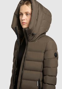 Brun pufferjakke med en stor hætte, der har lynlås lukning, sidelommer og et rund logo mærkat på venstre skulder. Blød, quiltet tekstur.