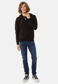 Sudadera negra con cremallera y capucha ajustable, combinada con jeans azules y zapatillas grises. El tejido parece suave y tiene una textura lisa.