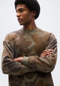 Νεαρός άνδρας με σταυρωμένα τα χέρια, φοράει μπλούζα μακρυμάνικη με καμουφλαρισμένο καφέ και πράσινο μοτίβο, κοιτά προς τα αριστερά του, μπροστά σε ένα απλό φόντο.