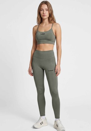 Frau steht in olivgrünem Sport-BH und Leggings mit "hummel" Logo, trägt weiße Sneakers, auf einem einfarbigen hellgrauen Hintergrund.