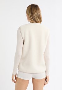 Světlé béžové fleecové tílko s texturovaným povrchem. Má zaoblený výstřih a hladný lem, nošené přes světlou dlouhý rukáv.