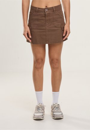 Hnedá mini sukňa s prednými vreckami a gombíkovými detailmi, nosená s bielymi ponožkami a masívnymi bielymi teniskami, stojaca na jednoliatom pozadí.