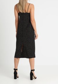 Svart midjelång klänning med färgglada prickar, tunna axelband och slits baktill. Materialet verkar lätt och strukturerat. Klackarna är svarta med remmar runt vristen.