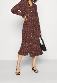Donna che indossa un abito midi a righe rosse e nere con bottoni sul davanti, tiene in mano una borsa nera arricciata e indossa sandali neri con tacco.