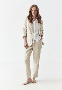Lněný oblek v světle béžové barvě s tailored sakem a odpovídajícími kalhotami; pod tím bílá košile s knoflíky. Neformální béžové pantofle.