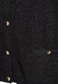 Melna teksturēta buklē auduma virsma ar trim spīdīgiem zelta pogām vienā pusē un vienu zelta pogu mazā horizontālā kabatas vāciņā.