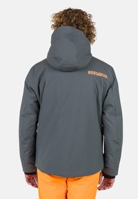 Graue isolierte Jacke mit Kapuze, die ein auffälliges oranges "Rossignol"-Logo auf dem Rücken hat. Glatte Textur, entworfen für den Außeneinsatz.