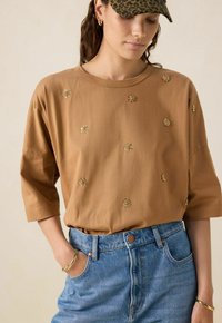 Chemise brune à manches courtes en coton avec broderie florale dorée, associée à un short en denim bleu taille haute. Coupe décontractée avec un col rond.