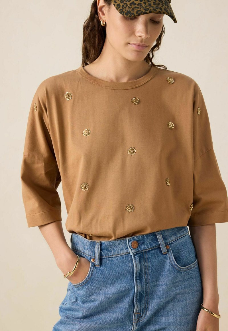 Chemise brune à manches courtes en coton avec broderie florale dorée, associée à un short en denim bleu taille haute. Coupe décontractée avec un col rond.