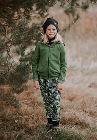 Zielona bluza z zamkiem, zielono-czarne spodnie dresowe w tropikalne liście, do tego czarna czapka i szare buty.